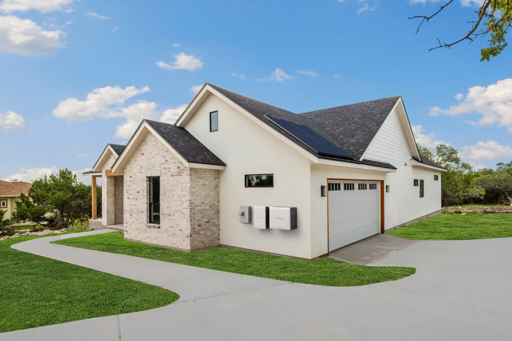 Modern light gray, single-story house with a two-car garage, featuring dark solar panels installed on the roof and wall-mounted battery units on the side exterior under a clear blue sky.